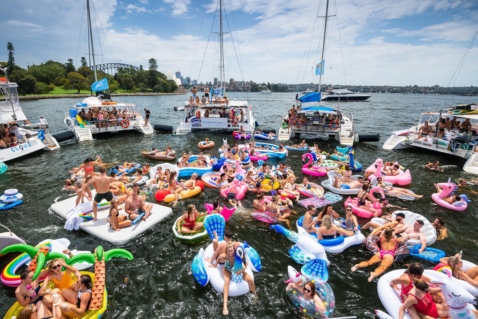 A lively scene of numerous people enjoying a summer day on a lake, with many on colorful inflatables in the water. Several boats are docked nearby, bustling with energy reminiscent of The Yacht Social Club Sydney Boat Hire events. The sky is mostly clear, and the background features trees and a distant cityscape.