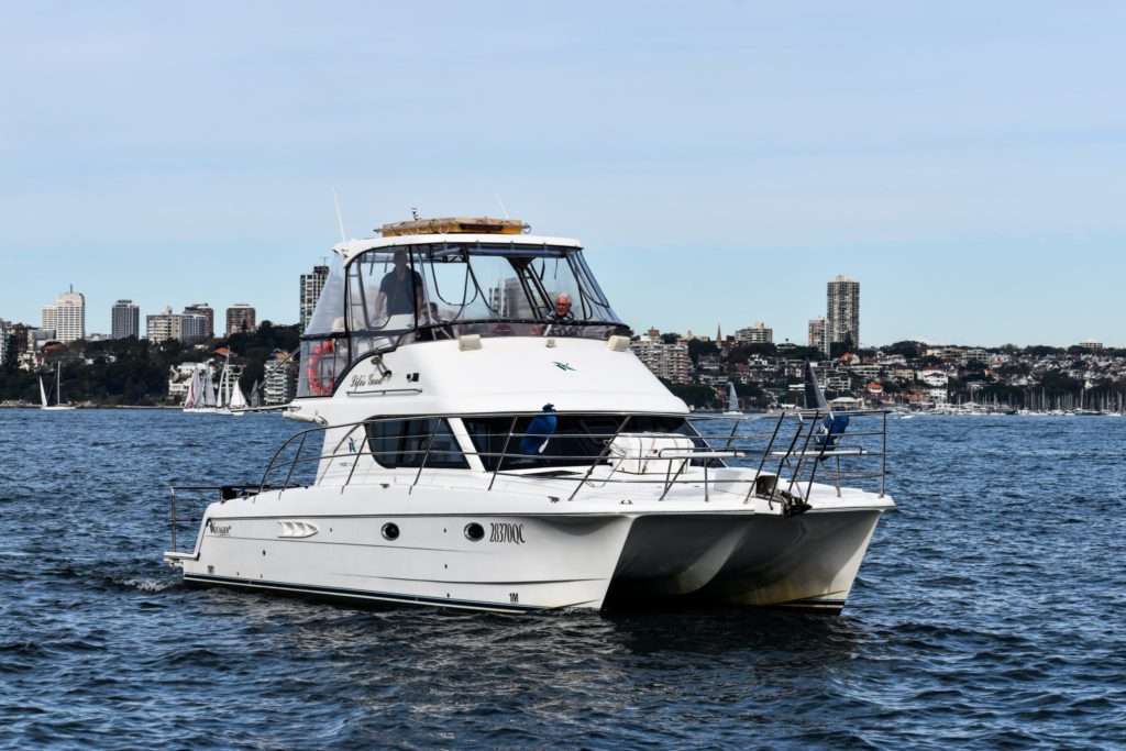 A white catamaran yacht sails on calm water with a city skyline and several smaller boats in the background, perfect for a private yacht charter in Sydney Harbour.