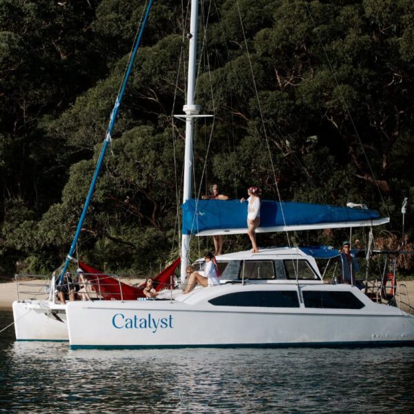 The white catamaran Catalyst is anchored by a forested shore. People are relaxing on deck, with one standing atop the boat and others lounging in red hammocks at the back.