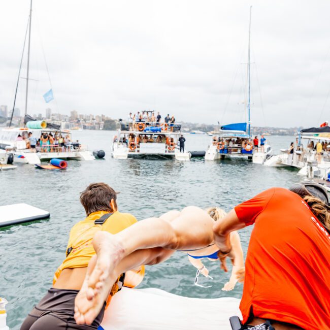 Three people jump off a boat into the water during a lively Yacht Social Club party. Multiple boats with revelers are seen in the background on a cloudy day. The mood is festive, with people enjoying the event surrounded by inflatables and water sports equipment.