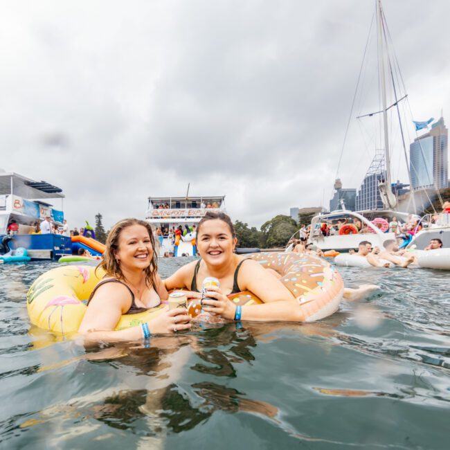 Two women in pool floats holding drinks smile at the camera while floating in harbor water. They are among several people on floats near boats, with a city skyline and cloudy sky in the background. The lively scene is reminiscent of The Yacht Social Club's event boat charters, adding an air of festivity.