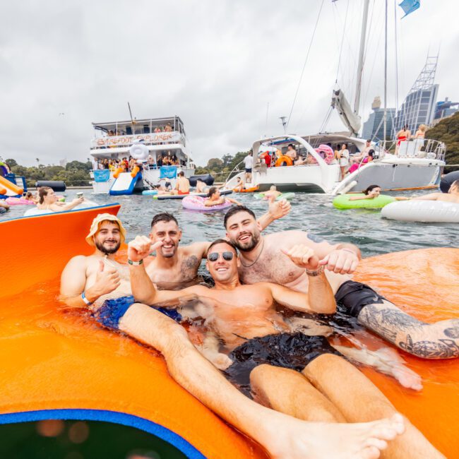 A group of four men smiles and poses for a photo on an orange float in the water. They are surrounded by others enjoying the water on inflatables and boats in the background. The sky is cloudy, and city buildings are visible in the distance, capturing a moment at The Yacht Social Club Sydney Boat Hire.
