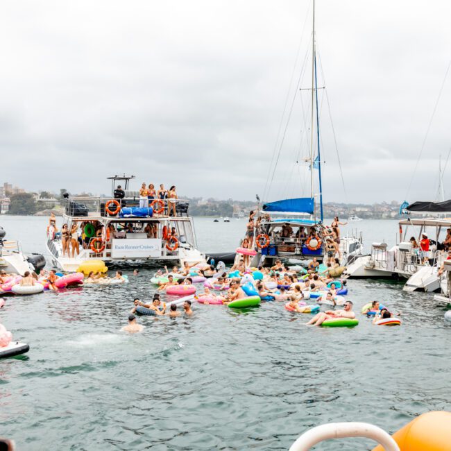 A lively scene on the water with numerous people on inflatable floats between several anchored boats. Participants of The Yacht Social Club are enjoying a social gathering, with music and activities in an overcast setting. Cityscape in the background.