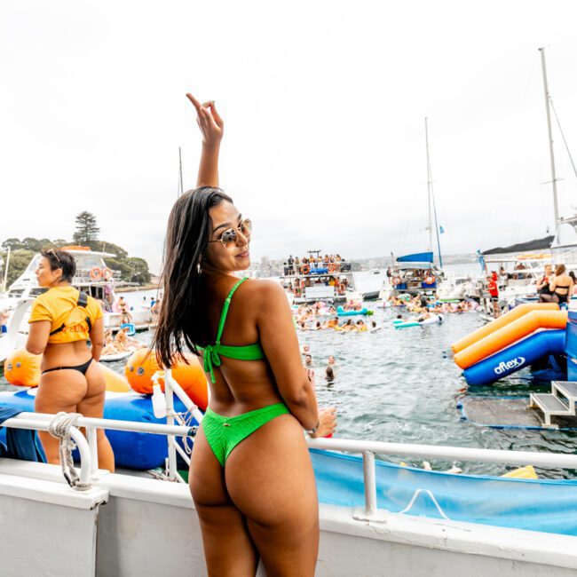 A woman in a bright green swimsuit, facing away from the camera with one arm raised, stands at the railing of a boat. She is smiling and looking back. In the background, people enjoy a festive atmosphere on multiple boats and inflatables in the water, all part of The Yacht Social Club Sydney Boat Hire event.