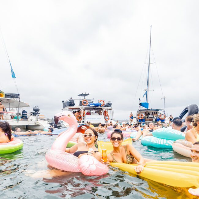 A lively scene at The Yacht Social Club event features many people enjoying the water on various inflatable floats, including a large pink flamingo. The sky is overcast. Several boats are anchored in the background, and guests are socializing and having fun.
