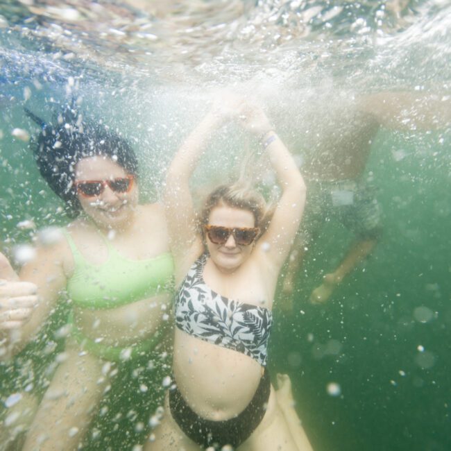 Two people underwater in a pool, wearing swimsuits and sunglasses. One in a green bikini top gives a thumbs-up, while the other in a patterned swimsuit top raises their arms. Bubbles and water movement surround them. "The Yacht Social Club Event Boat Charters" logo is in the bottom right corner.