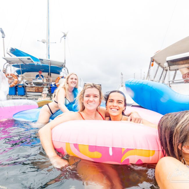 Three smiling women in swimsuits are floating on inflatable tubes, with a boat from The Yacht Social Club in the background. The scene is bright and cheerful, suggesting they're enjoying a day on the water. More inflatable toys and another boat are also visible, capturing the joyous vibe of Boat Parties Sydney.