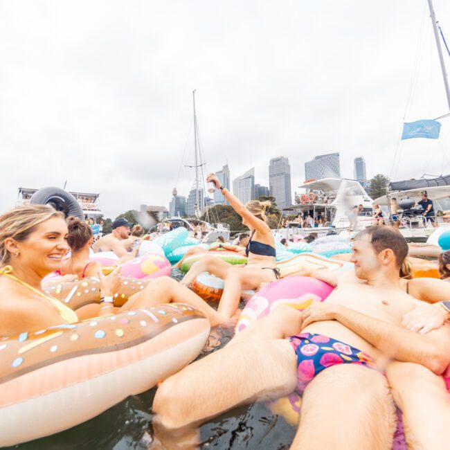 A lively scene of people enjoying a day on the water, floating on colorful inflatable tubes shaped like donuts. The background features a city skyline with high-rise buildings, and the event appears to be happening near several anchored boats from The Yacht Social Club.