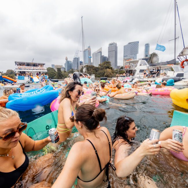 A lively scene of people enjoying a pool party on a cloudy day. They are in an inflatable pool on a boat, raising drinks and socializing. Various pool floats and nearby yachts from The Yacht Social Club are visible, with a city skyline in the background.