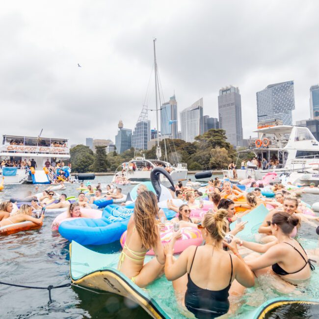 A large group of people enjoy a party on inflatable floats in the water, surrounded by several boats. They are near a city skyline with tall buildings under a cloudy sky. Everyone appears to be having fun. The atmosphere is lively and festive, reminiscent of Boat Parties Sydney The Yacht Social Club.