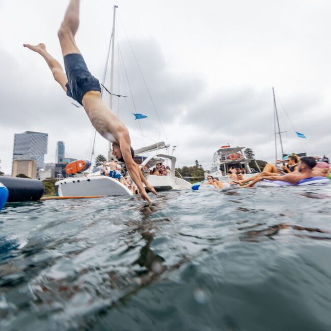 A man in mid-air dives into the water surrounded by boats and people on inflatable floats. The city skyline and overcast sky are visible in the background. The scene is lively with several people enjoying the water, chatting on boats from Sydney Harbour Boat Hire, and partying at The Yacht Social Club.