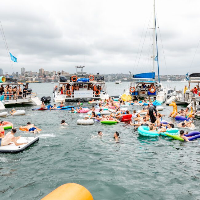 A lively scene of people swimming and floating on various inflatable rafts and tubes in a body of water, surrounded by several anchored boats. It appears to be a festive gathering organized by The Yacht Social Club Event Boat Charters, with the skyline of a city visible in the background. The sky is overcast.