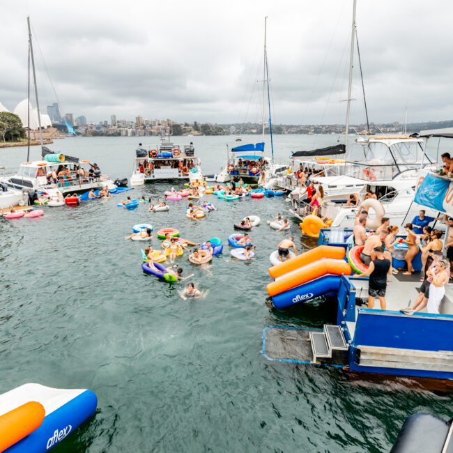 A bustling scene of people enjoying a water party with multiple boats anchored close together. People are swimming, lounging on inflatables, and socializing on the boats. The Yacht Social Club Sydney Boat Hire offers a stunning backdrop of a city skyline under a cloudy sky.
