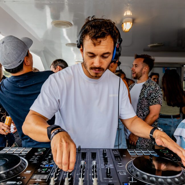 A DJ wearing a white t-shirt and headphones is intently working on a mixing console aboard a boat. Several people are around, some chatting and enjoying drinks. The atmosphere is lively and casual, suggesting a party by The Yacht Social Club Event Boat Charters in Sydney.