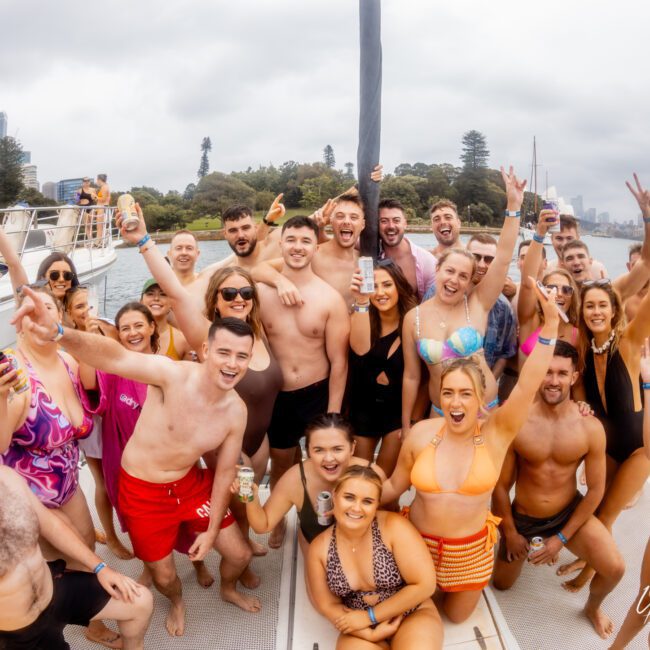 A group of 23 people enjoying The Yacht Social Club Sydney Boat Hire on a boat. Many are in swimwear, smiling, and holding drinks. The background shows a scenic view with water, trees, and city buildings under a cloudy sky. The vibe is festive and cheerful.