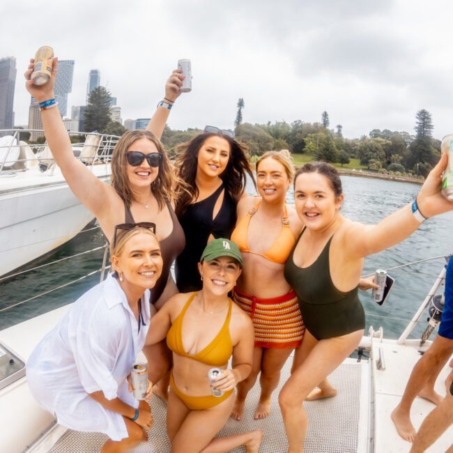 A group of six women stands on a boat's deck, smiling and raising their drinks in celebration during a Boat Parties Sydney The Yacht Social Club event. They are wearing swimsuits and casual summer clothing. The background features a glimpse of city buildings and trees under a cloudy sky.