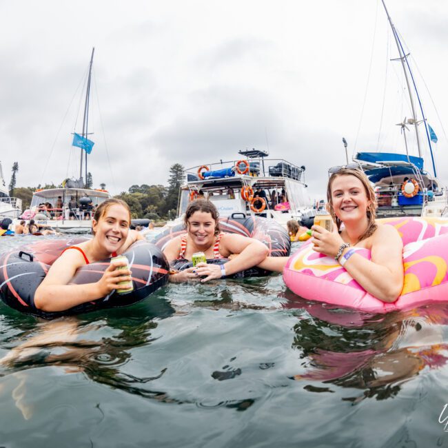 Three women in colorful inflatable tubes floating on the water, holding drinks and smiling at a yacht social event by Boat Parties Sydney The Yacht Social Club. Boats and yachts are visible in the background on a cloudy day.