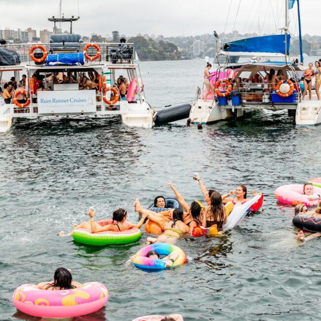 A lively group of people enjoy a party in the water, floating on colorful inflatables near two anchored yachts. The yachts, part of The Yacht Social Club Sydney Boat Hire, have passengers onboard, and there's a cityscape in the background under a cloudy sky. The atmosphere is festive and cheerful.