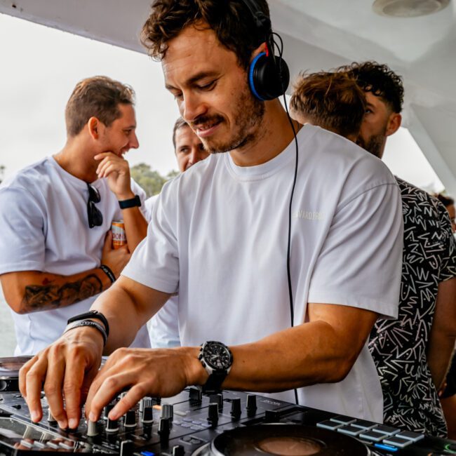 A man wearing a white T-shirt and headphones is DJing on a boat from The Yacht Social Club Sydney Boat Hire, smiling as he operates the mixer. Several people are gathered around him, one in a patterned shirt and others in casual outfits. The scene appears relaxed and social.