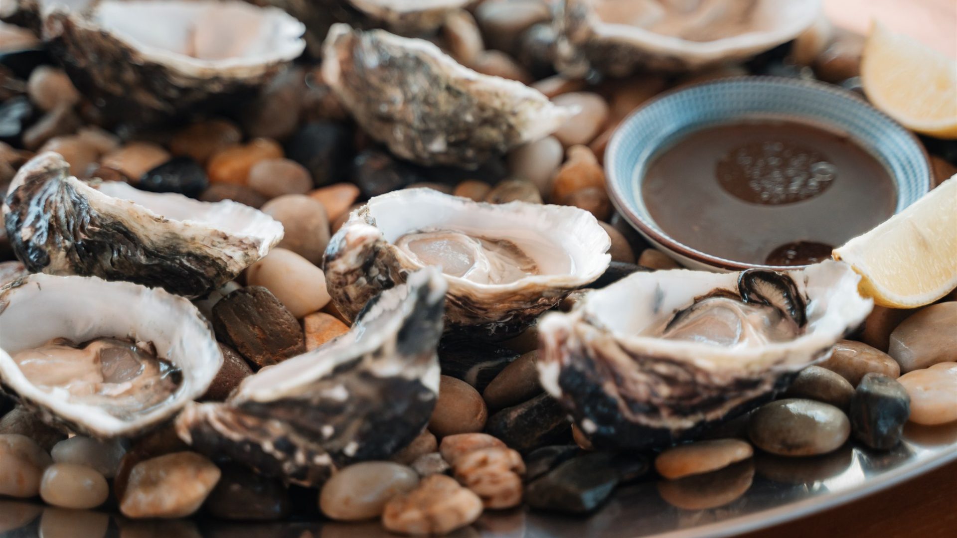 A plate of opened oysters on the half shell, resting on a bed of mixed small stones. A small bowl of brown sauce and some lemon wedges are placed beside the oysters.