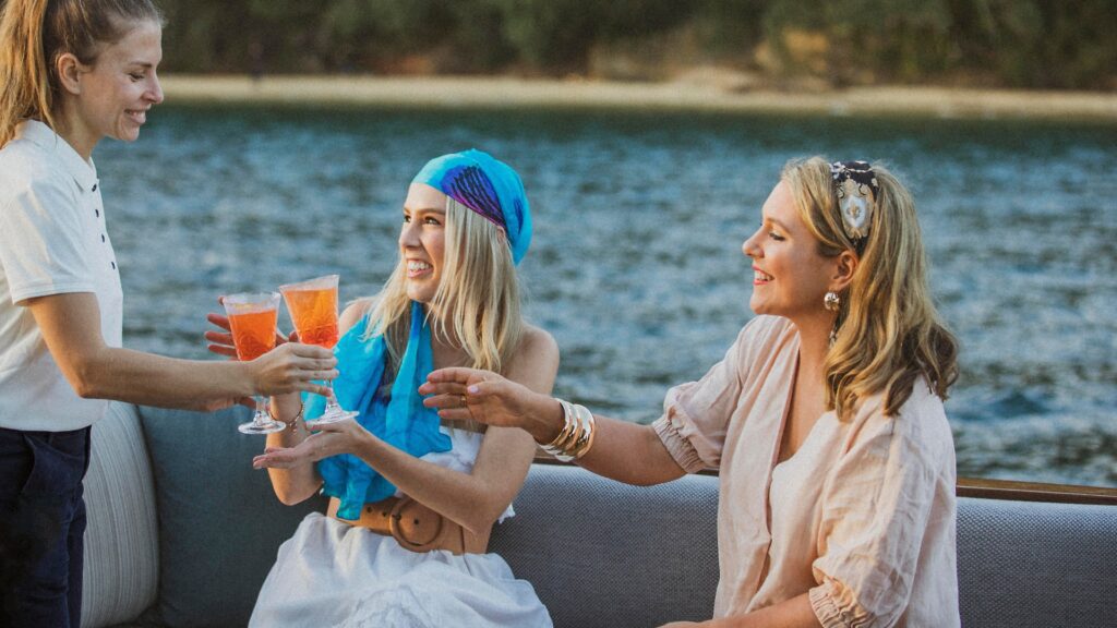 Two women sit on an outdoor couch by the water, smiling and reaching for cocktails served by a person in a white shirt. One woman wears a blue headscarf and white dress, while the other wears a light blouse. The scene is cheerful and relaxed.