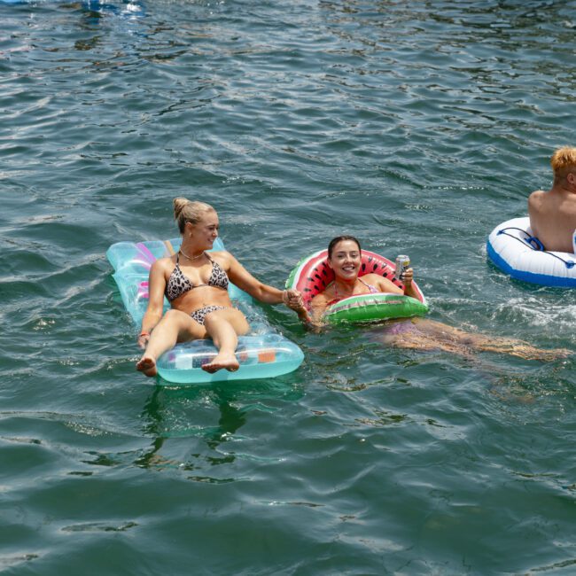 Two people lying on inflatable pool floats, smiling and relaxing in a large body of water. One person on a colorful float and the other on a watermelon-themed one. A third person is in the background on a blue and white float.