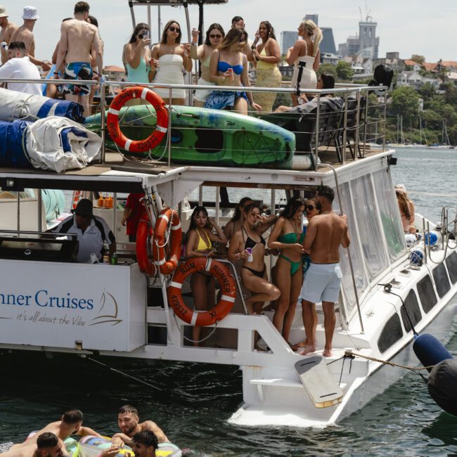 A crowded boat party with people in swimsuits enjoying music and drinks. Some are lounging on the boat's upper deck, while others swim in the water nearby. Bright life rings are visible on the boat. Buildings are in the distant background.