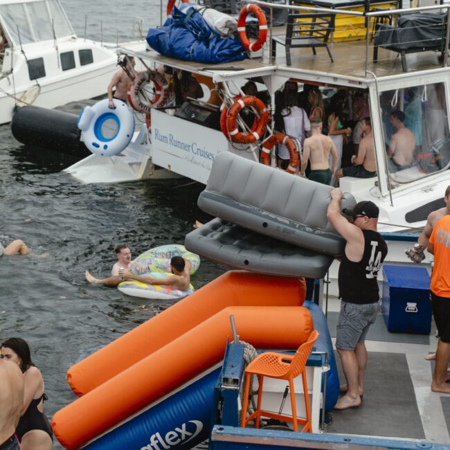 People enjoy a party on a docked boat. A person carries inflatables, while others swim and relax near the boat. The scene is lively with floats and life rings visible. The water is calm, and the weather appears overcast.