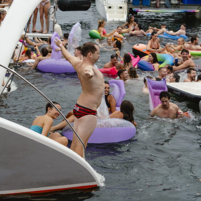People enjoying a lively pool party on the water, surrounded by inflatable floats. A person in swim trunks is jumping into the water from a yacht, while others relax or swim nearby. A boat is visible in the background.