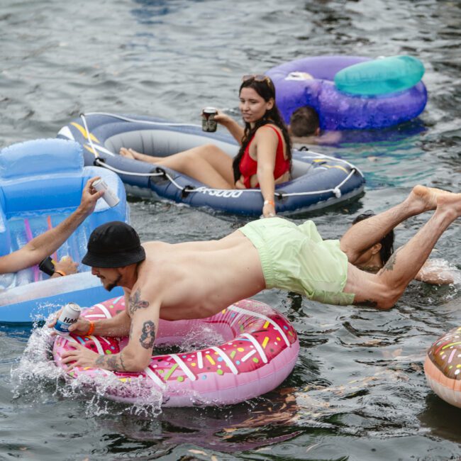 A group of people enjoying a sunny day on a lake, floating on colorful inflatables. A man in green shorts is diving onto a pink donut-shaped float, while a woman lounges on a blue and purple inflatable nearby, holding a drink.