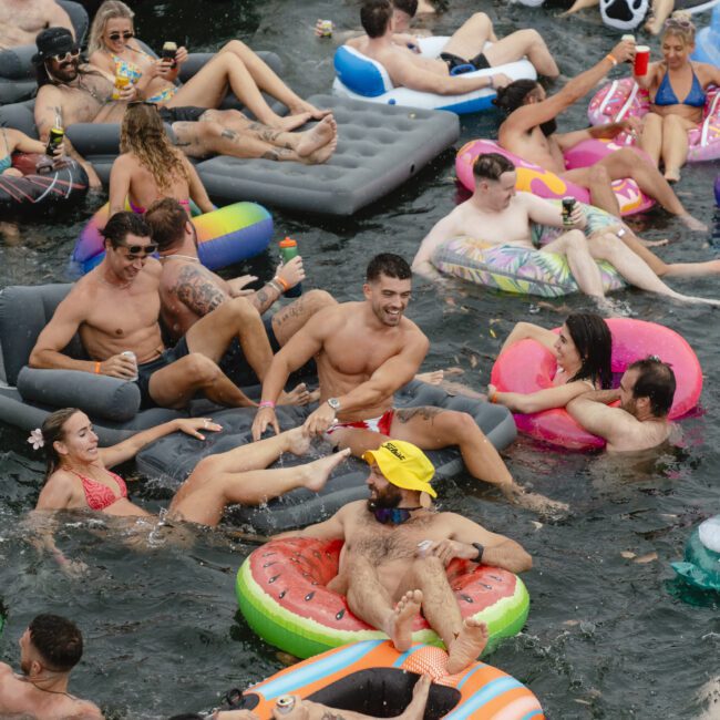 A group of people relax on inflatable floats in the water, wearing swimwear. The scene is lively, with colorful floats shaped like pizza and watermelon. Everyone is enjoying the sunny day and drinks.