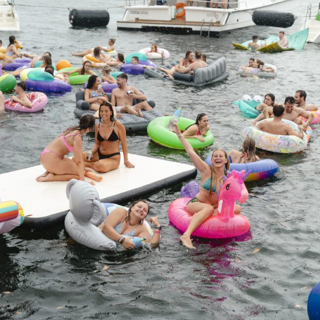 A lively scene on a lake with people enjoying a floating party. Adults relax on colorful inflatables, including animals and unicorns, near a docked boat. The city skyline is visible in the background under a cloudy sky.
