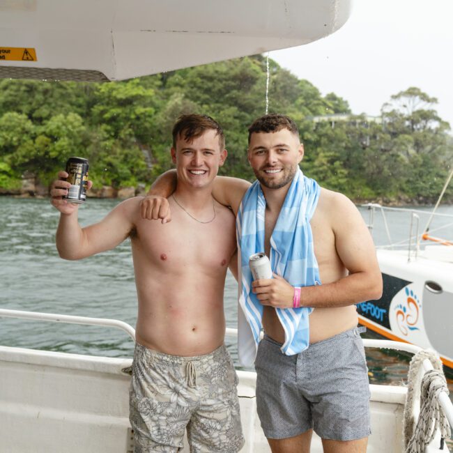 Two men smiling and holding drinks on a boat. They're shirtless, with one wearing patterned shorts and the other in gray shorts with a blue towel around his shoulders. Trees and water are in the background.