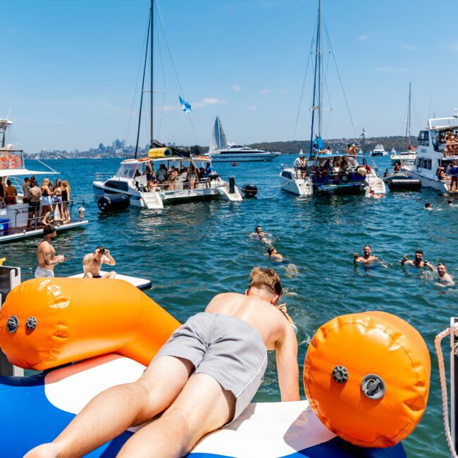 A vibrant scene of people enjoying a yacht party on a sunny day. Several boats are anchored, with guests swimming and socializing. A person relaxes on an inflatable float in the foreground. The city skyline is visible in the distance.