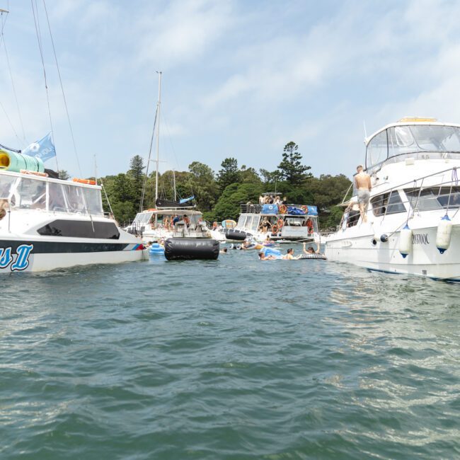 Boats anchored in calm water with people relaxing on rafts and swimming. Trees and a partly cloudy sky in the background.