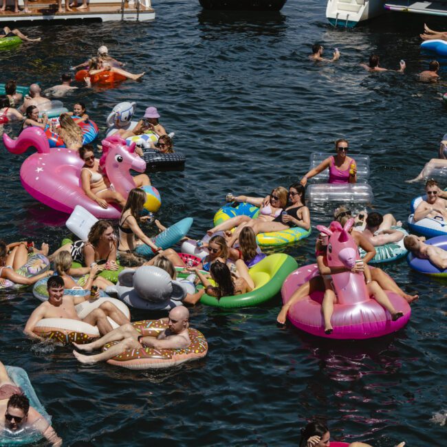 A large group of people floating in a lake on various inflatable rafts, including unicorns, donuts, and flamingos. The crowd is lively and enjoying the sunny day. Boats are visible in the background.