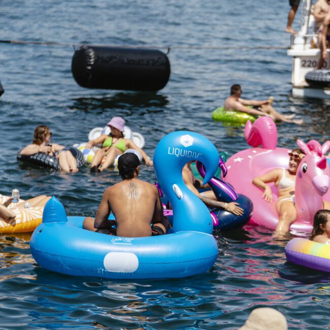 People are floating on inflatable pool toys, including a blue swan and a pink unicorn, in a body of water. There are several others relaxing nearby, and a boat is visible in the background.