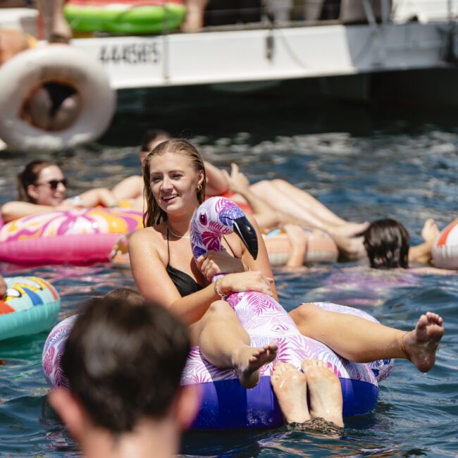 A woman in a black swimsuit is smiling and sitting on a pink inflatable flamingo in the water, surrounded by people on various colorful inflatables. A boat is in the background. The scene is lively and sunny.