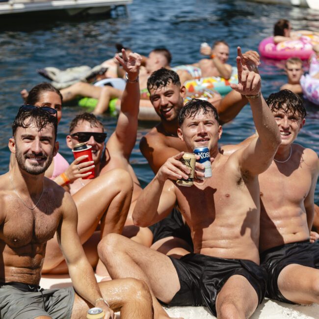 A group of shirtless men and other people enjoy a sunny day on the water, sitting and floating on inflatable tubes. They are smiling, holding drinks, and appear relaxed and happy. The water and inflatables create a vibrant and fun atmosphere.