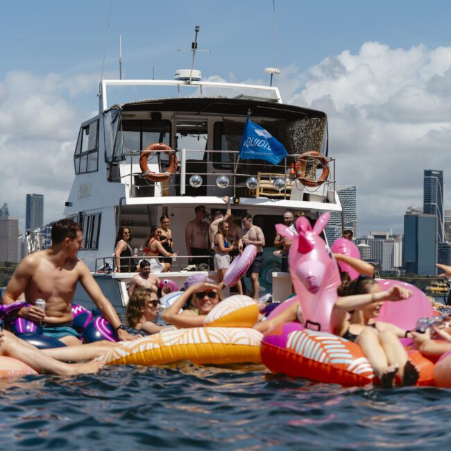 A group of people relax on colorful inflatable pool floats in the water near a boat. In the background, a city skyline is visible under a partly cloudy sky. Some people wave and enjoy sunny weather.