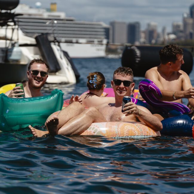 A group of people relaxing on colorful inflatables in the water, smiling and holding drinks. Boats and a city skyline are visible in the background. It's a sunny day and everyone seems to be enjoying the scene.