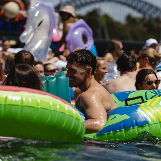 A group of people enjoying a sunny day in the water with colorful inflatable pool floats, including a large green one in the foreground. The setting is lively, with a bridge visible in the background.