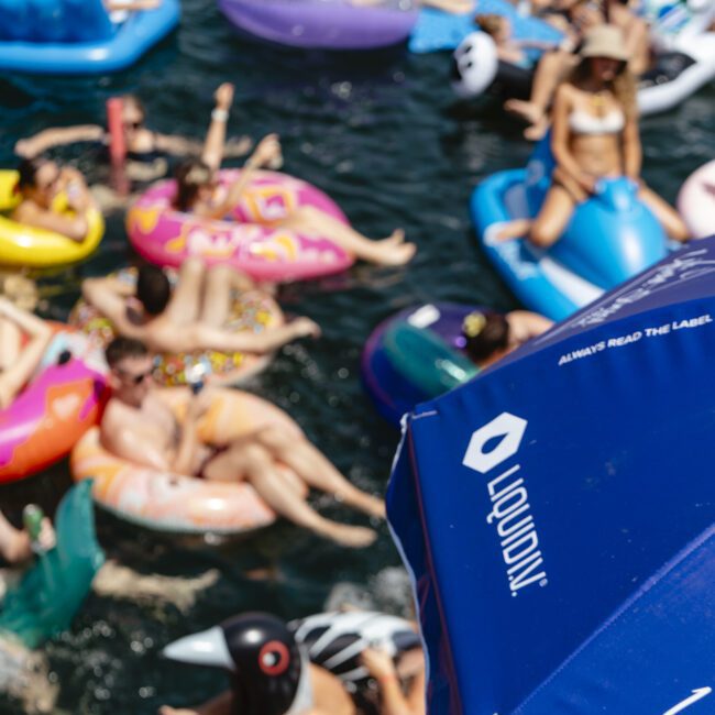 A crowded scene on a lake with people relaxing on colorful inflatable rafts. A blue umbrella is in the foreground, partially covering the view. Various float shapes are visible, including birds and animals.