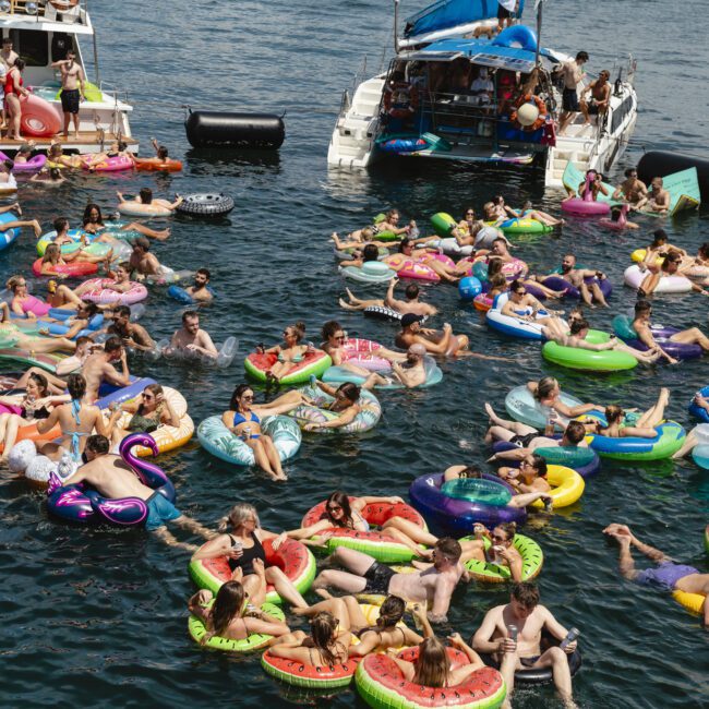 A lively scene of numerous people floating on colorful inflatable pool floats in a body of water, surrounded by two boats. Floats include shapes like donuts, flamingos, and pizza slices. People are enjoying the sunny day, socializing and relaxing.