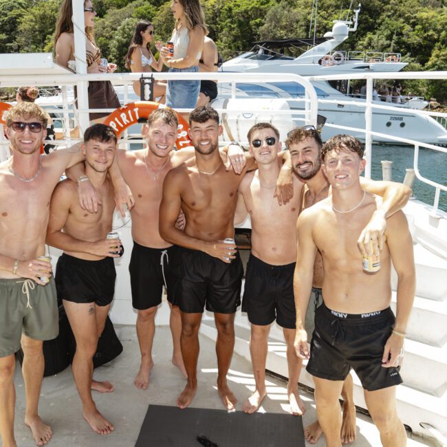 A group of seven young men in swimsuits stand together on a boat deck, smiling with arms around each other. People are in the background on the upper deck, with a yacht and trees visible in the distance.