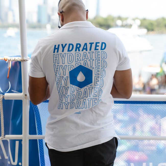 A man stands with his back to the camera, wearing a white shirt with the word "HYDRATED" repeated along with a water droplet graphic. He appears to be on a boat or dock with water and a city skyline in the background.