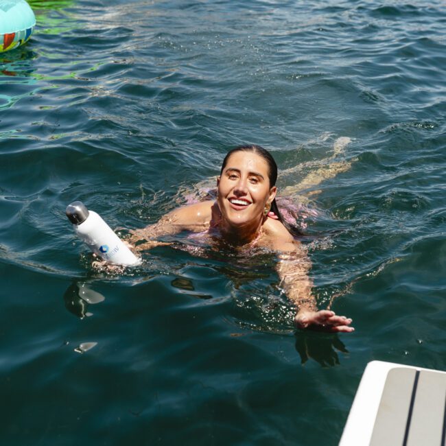 A person swimming in a lake smiles while holding a water bottle. The water is dark and reflective. A colorful inflatable ring is visible in the background. Part of a dock is seen in the foreground.