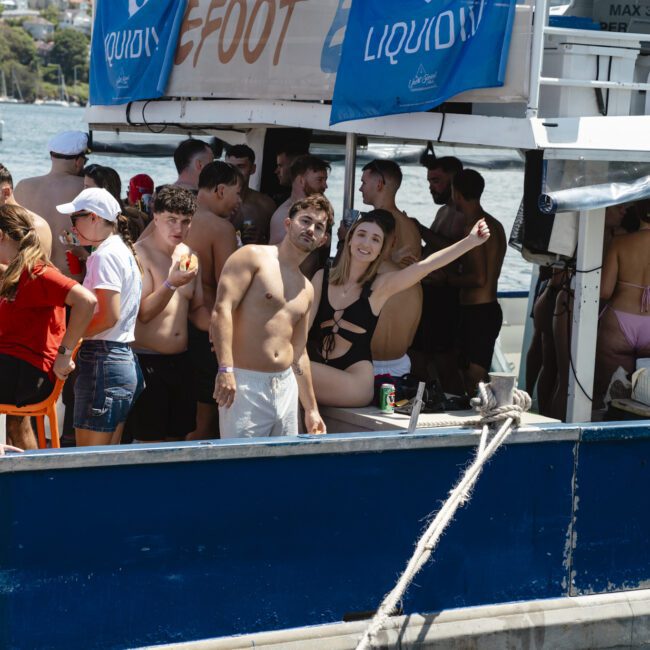 A group of people in swimwear are enjoying a party on a boat. Some are dancing and holding drinks, while others chat. The boat is docked near a scenic waterfront, with trees and sailboats in the background.