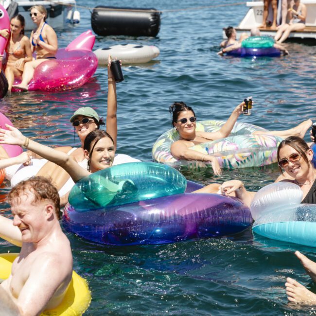 A group of people relaxing on colorful inflatable rings in a lake, holding drinks and smiling. They are surrounded by inflatables shaped like a flamingo and a unicorn. The sun is shining, creating a festive and joyful atmosphere.