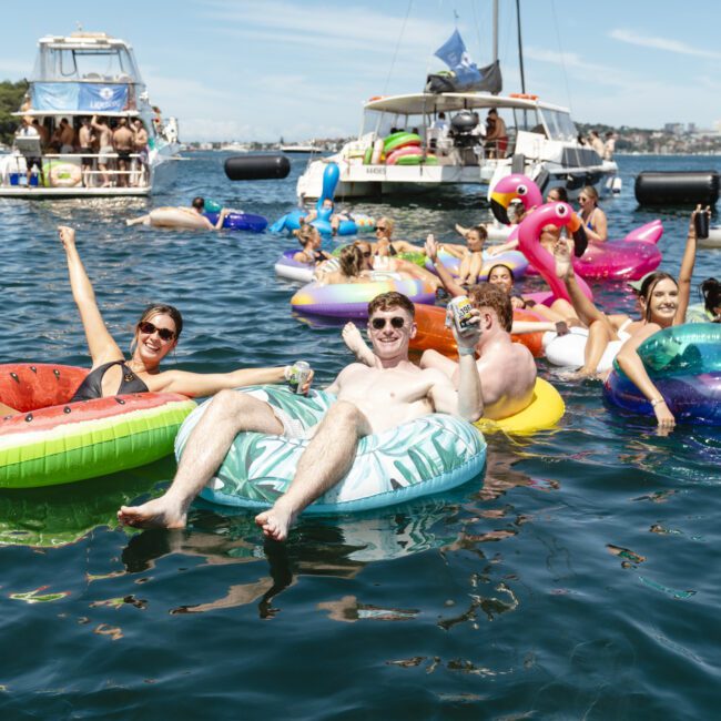 A group of people enjoying a sunny day on the water, lounging on colorful inflatable floats, including a unicorn and a flamingo. Boats are in the background, and everyone looks relaxed and cheerful.